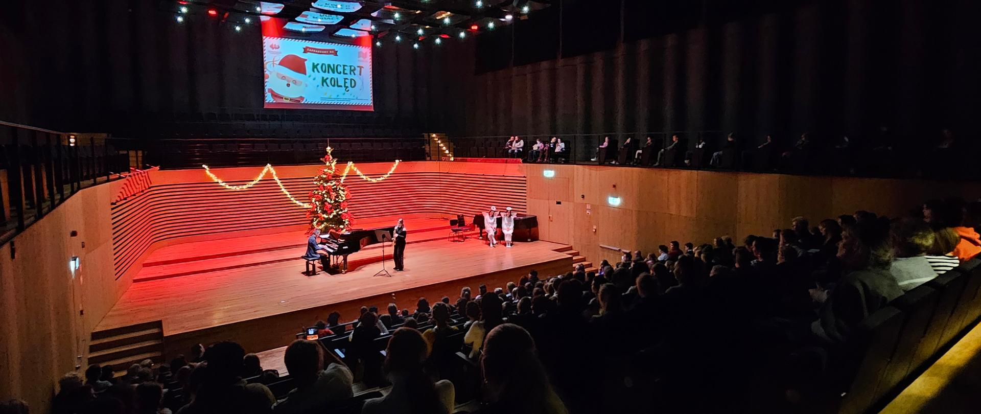 Nastolatka stojąc na środku estrady gra na saksofonie, za nią na fortepianie gra mężczyzna. Po prawej stronie dwie kobiety w białych strojach aniołków siedzą na krzesłach, z tyłu udekorowana choinka. Z przodu od tyłu widać widownię z publicznością.