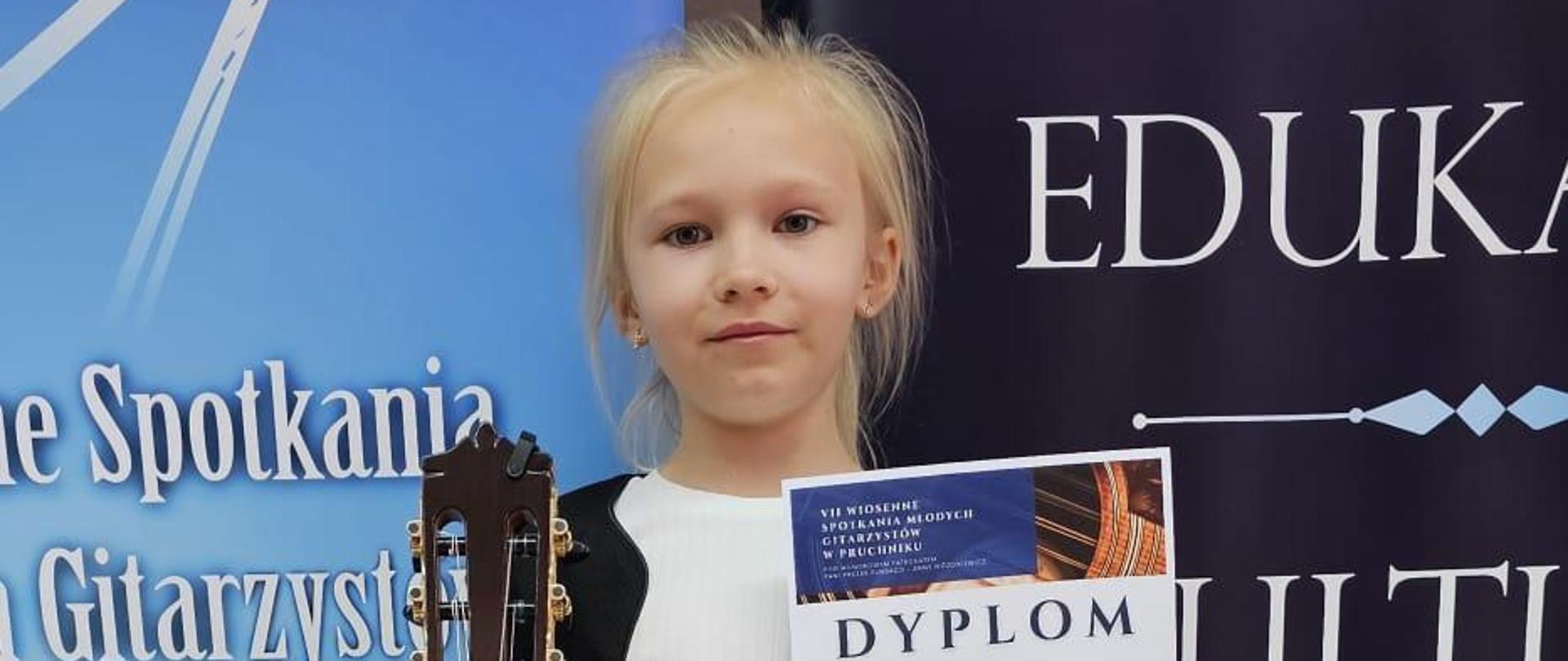 Zuzanna Stach - VII Wiosenne Spotkania Młodych Gitarzystów w Pruchniku. Uczennica z gitarą i dyplomem, w tle reklama konkursu.