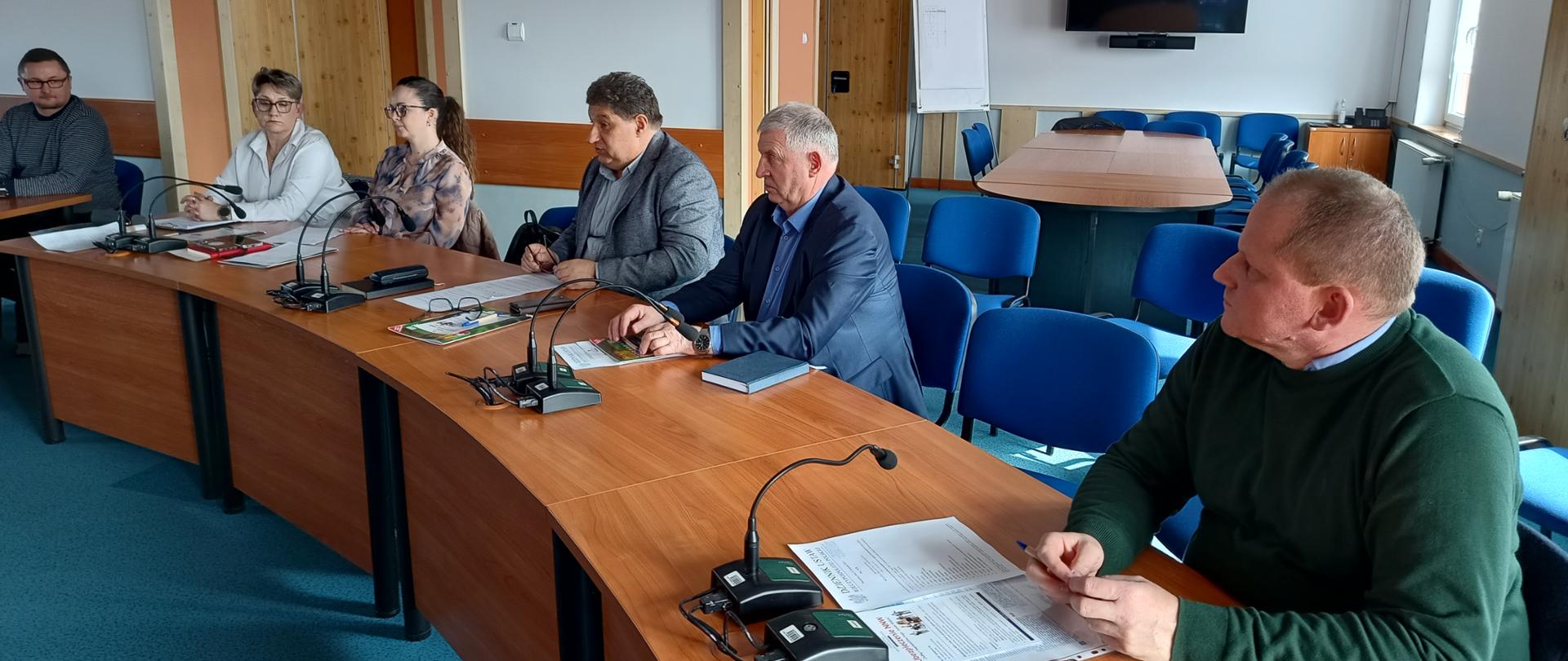 Sala konferencyjna z długimi, drewnianymi stołami ustawionymi w kształt podkowy. Po lewej stronie siedzi kilka osób – kobiety i mężczyźni w średnim wieku – ubranych w stylu biznesowym i półformalnym. Na stołach znajdują się mikrofony konferencyjne, dokumenty, notesy oraz długopisy. Jedna z osób trzyma kartki, inna ma przed sobą zamknięty notatnik. W tle widać drzwi, ściany w jasnych kolorach oraz dodatkowe krzesła ustawione pod ścianą. Atmosfera sugeruje formalne spotkanie lub naradę.