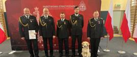 Strażacy w mundurach wyjściowych pozują do zdjęcia w tle bordowy baner na napisem Państwowa Straż Pożarna. Przy nodze jednego ze strażaków siedzi pies