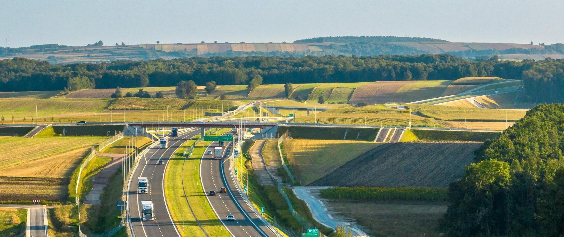 Dwujezdniowa droga ekspresowa S7 po dwa pasy ruchu w obu kierunkach. Droga przebiega wśród pól uprawnych i lasów ciągnących się po horyzont.