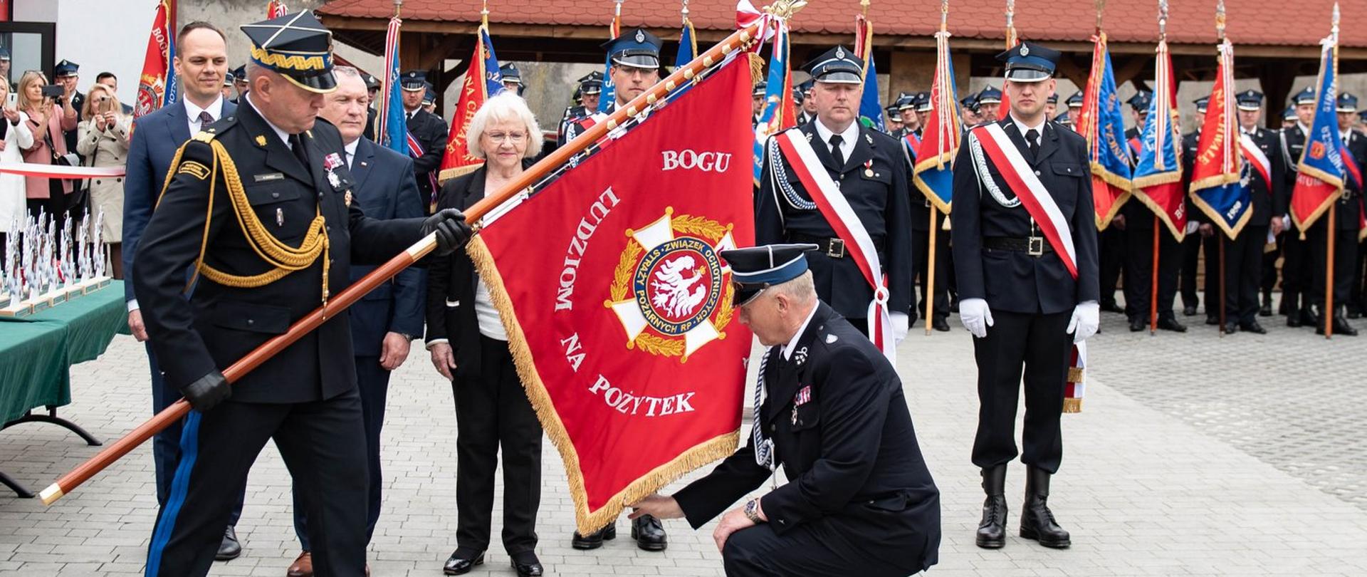 Zdjęcie zrobione na zewnątrz, podczas uroczystości. Po lewej stronie zdjęcia oficer Państwowej Straży Pożarnej w stopniu generała trzyma pochylony sztandar. Na sztandarze, na czerwonym tle umieszczono jest logo związku Ochotniczych Straży Pożarnych RP oraz widać część napisu "Bogu (...) Ludziom na pożytek". W tle stoją zaproszeni goście oraz poczty sztandarowe Ochotniczych Straży Pożarnych. 
