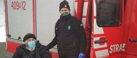 Na zdjęciu widać starszą osobę siedzącą na wózku inwalidzkim trzymającą w rękach kule ortopedyczne obok stoi strażak w ubraniu koszarowym. Osoby te mają zasłonięte usta i nos a za nimi znajduje się samochód ratowniczy straży pożarnej.