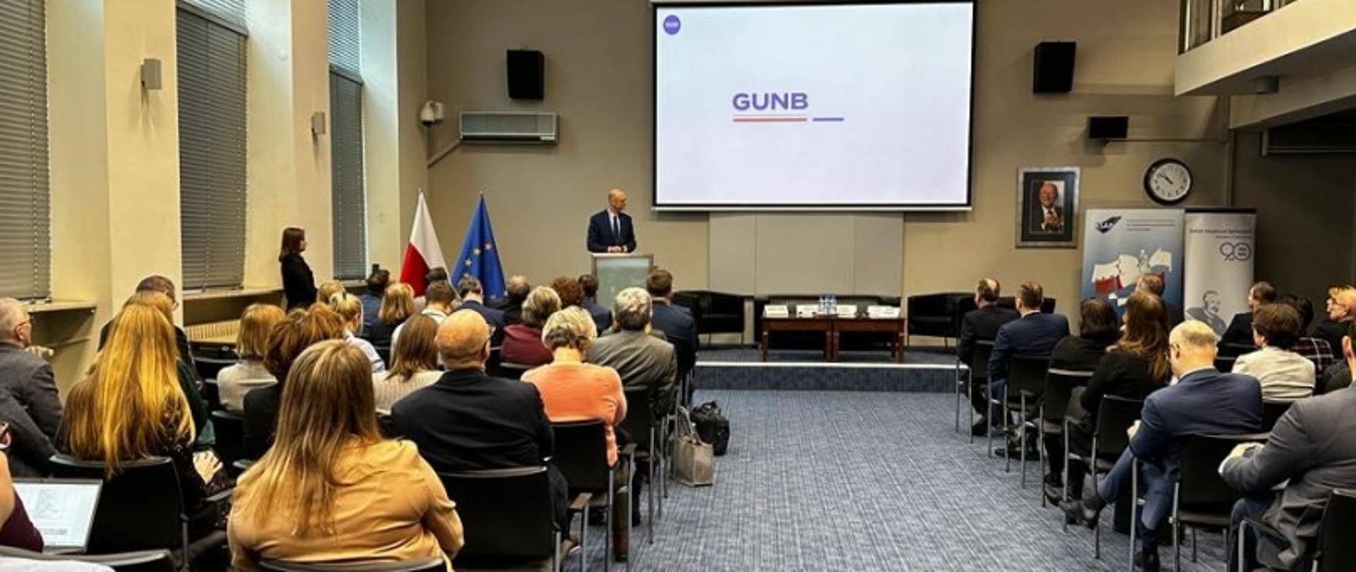 Prezentacja w sali konferencyjnej GUNB z prelegentem przy mównicy i słuchaczami.