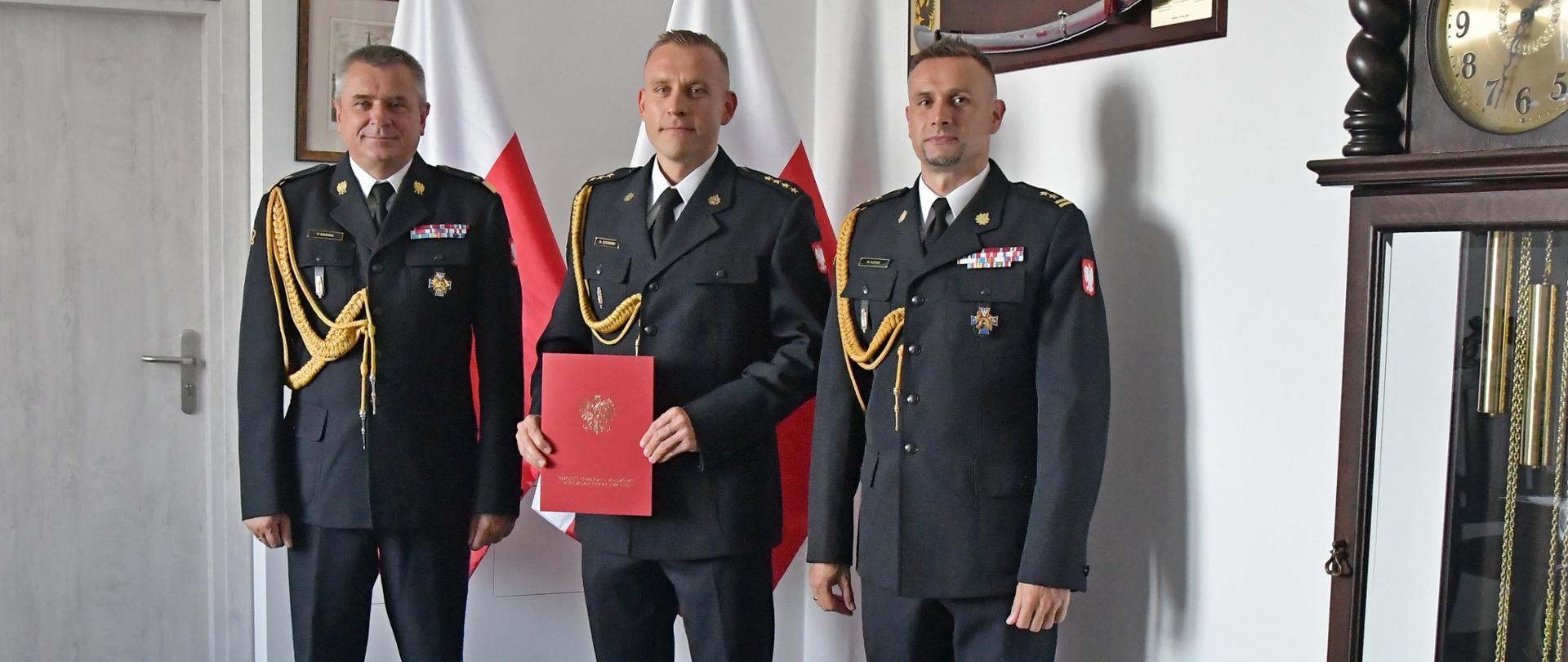 Trzech funkcjonariuszy Państwowej Straży Pożarnej stoi obok siebie strażak w środku trzyma czerwoną teczkę za nimi stoją dwie flagi Polski wisi obraz i szabla znajdują się drzwi oraz niedaleko ustawiony jest obraz.