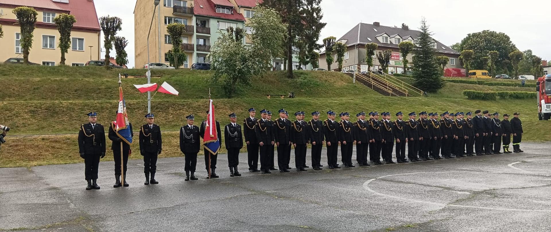 Na boisku w dwuszeregu stoją strażacy biorący udział w powiatowych obchodach Dnia Strażaka. Po lewej stronie stoją dwa poczty sztandarowe. Za nimi na słupie fiszą flagi Rzeczpospolitej Polski. Po prawej stronie stoi wóz strażacki.