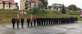 Na boisku w dwuszeregu stoją strażacy biorący udział w powiatowych obchodach Dnia Strażaka. Po lewej stronie stoją dwa poczty sztandarowe. Za nimi na słupie fiszą flagi Rzeczpospolitej Polski. Po prawej stronie stoi wóz strażacki.