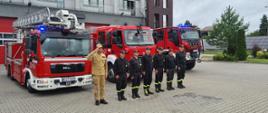 Na pierwszym planie strażacy ustawieni w szeregu, stojący na baczność. Oddają hołd w związku z Narodowym Świętem Dnia Pamięci Powstania Warszawskiego. Na drugim planie trzy samochody pożarnicze na tle budynku komendy.