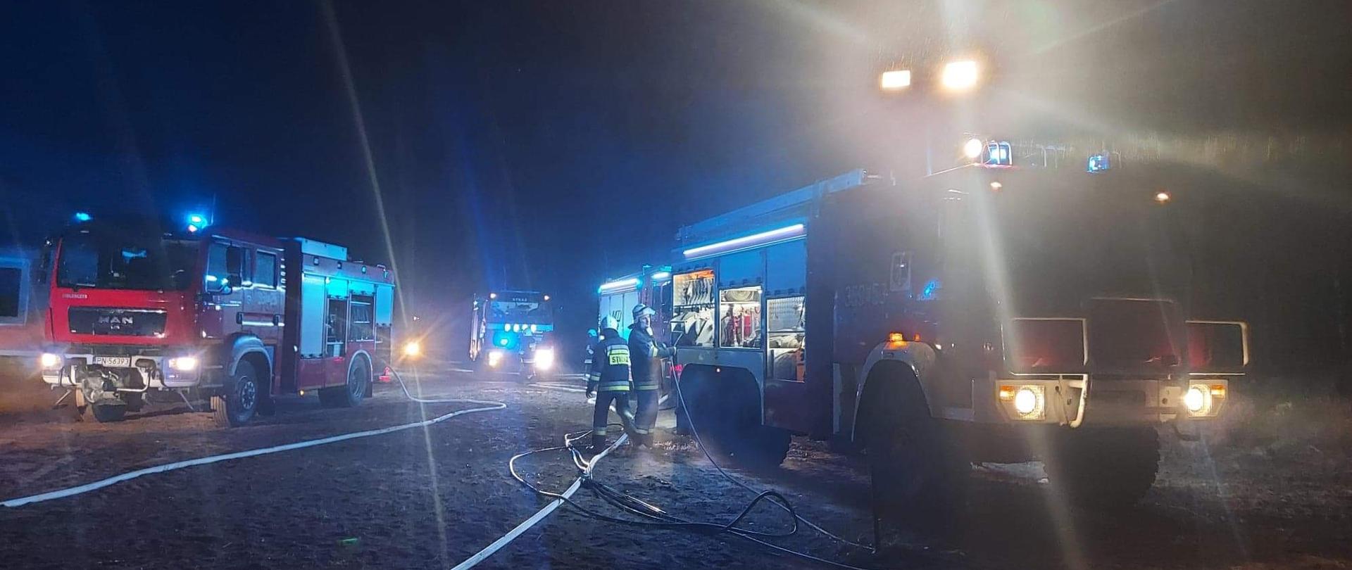 Zdjęcie przedstawia samochody strażackie oraz strażaków podczas gaszenie pożaru lasu. Samochody stoją w punkcie koncentracji sił i środków. 