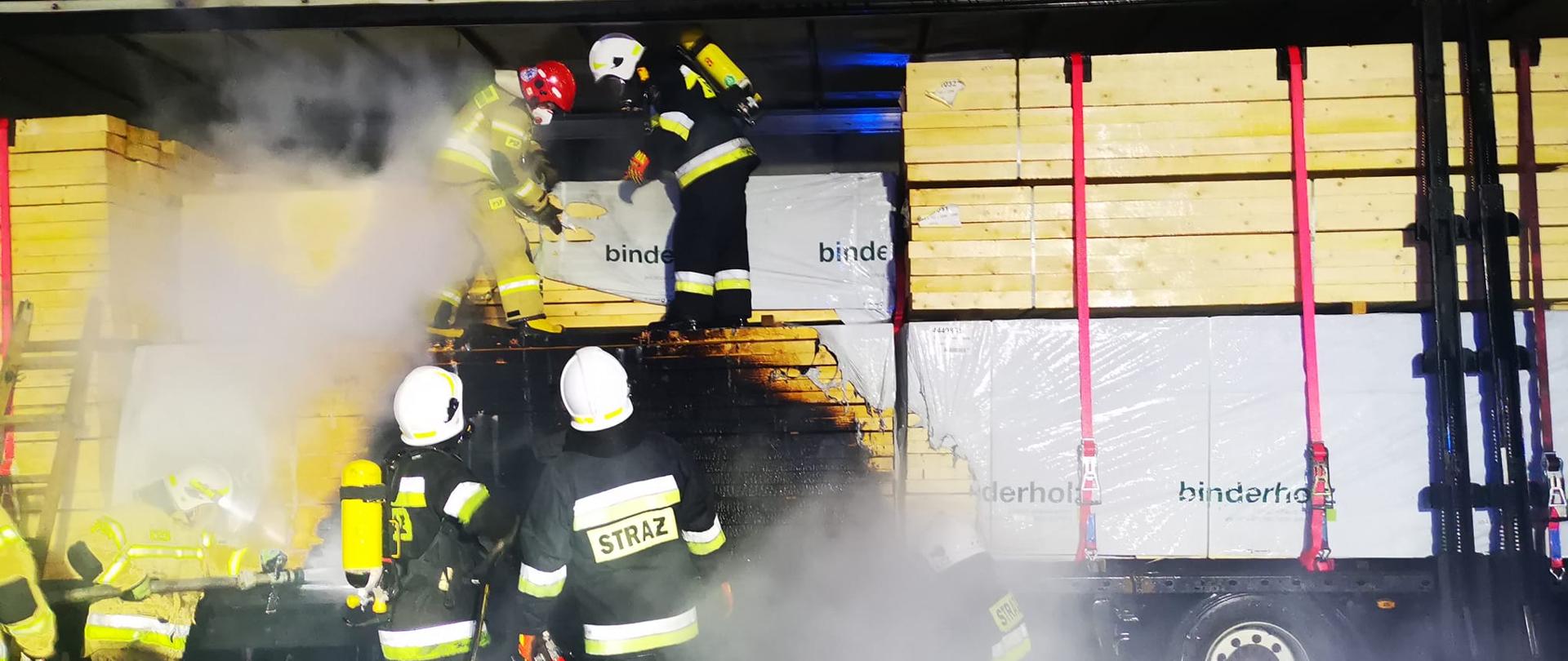 Na zdjęciu widać sześciu strażaków gaszących wnętrze naczepy załadowanej drewnem. Dwóch strażaków stoi na drewnie wewnątrz naczepy. Jeden ze strażaków ubrany jest w żółty strój i czerwony hełm a drugi w czarny strój i biały hełm. Na drodze stoją czterej strażacy. Dwóch ubranych jest w czarny strój i białe hełmy, trzeci w żółte ubranie i biały hełm a czwarty strażak ma żółty strój. O naczepę oparta jest drewniana drabina. Jest ciemno, ale teren jest oświetlony.
