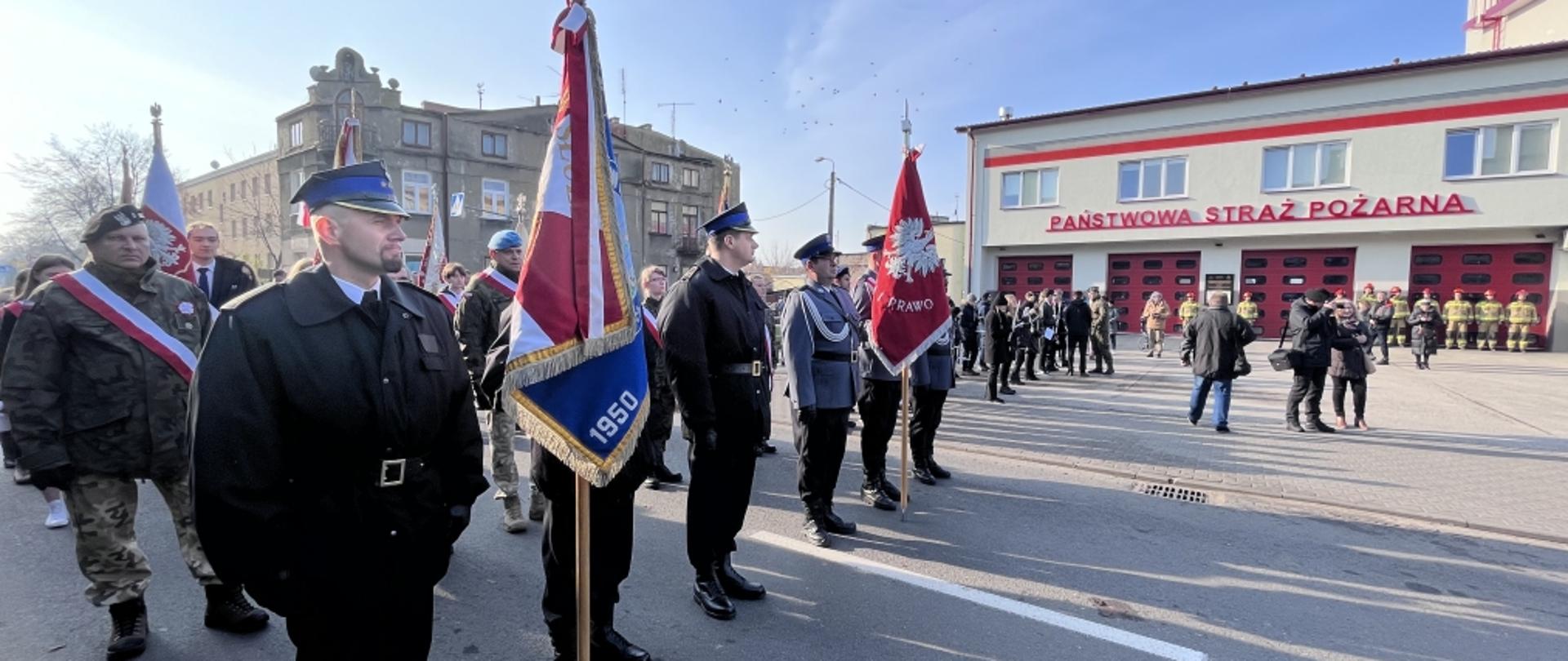 Na zdjęciu po lewej stronie widoczny poczet sztandarowy komendy powiatowej państwowej straży pożarnej. Funkcjonariusze w ubraniach wyjściowych, wysokich butach oraz kurtkach i rogatywkach. Po prawej stronie stoi poczet sztandarowy komendy powiatowej policji oraz widoczny jest budynek komendy powiatowej PSP, strażacy w ubraniach specjalnych stojący przed garażami i zebrani goście.