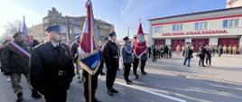 Na zdjęciu po lewej stronie widoczny poczet sztandarowy komendy powiatowej państwowej straży pożarnej. Funkcjonariusze w ubraniach wyjściowych, wysokich butach oraz kurtkach i rogatywkach. Po prawej stronie stoi poczet sztandarowy komendy powiatowej policji oraz widoczny jest budynek komendy powiatowej PSP, strażacy w ubraniach specjalnych stojący przed garażami i zebrani goście.