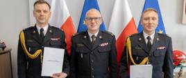 Pamiątkowe zdjęcie Zastępca KG PSP bryg. Pawł Frysztak, mł.bryg. Bogusław Szydło, mł.bryg. Dariusz Ruchała. Strażacy w umundurowaniu wyjściowym na tle flag państwowych i Unii Europejskiej