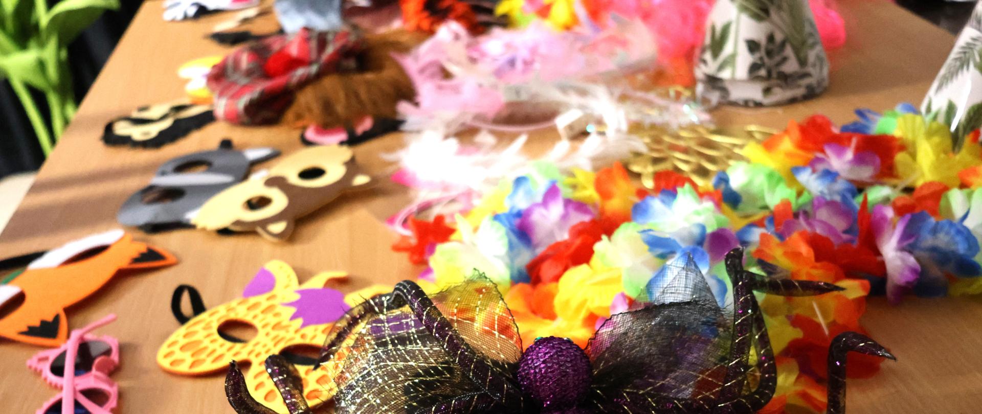 Na stoliku leżą kolorowe akcesoria, z przodu fioletowo czarna opaska w kształcie kokardy, dalej kolorowe okulary, kwiatowe naszyjniki, czapeczki papierowe.