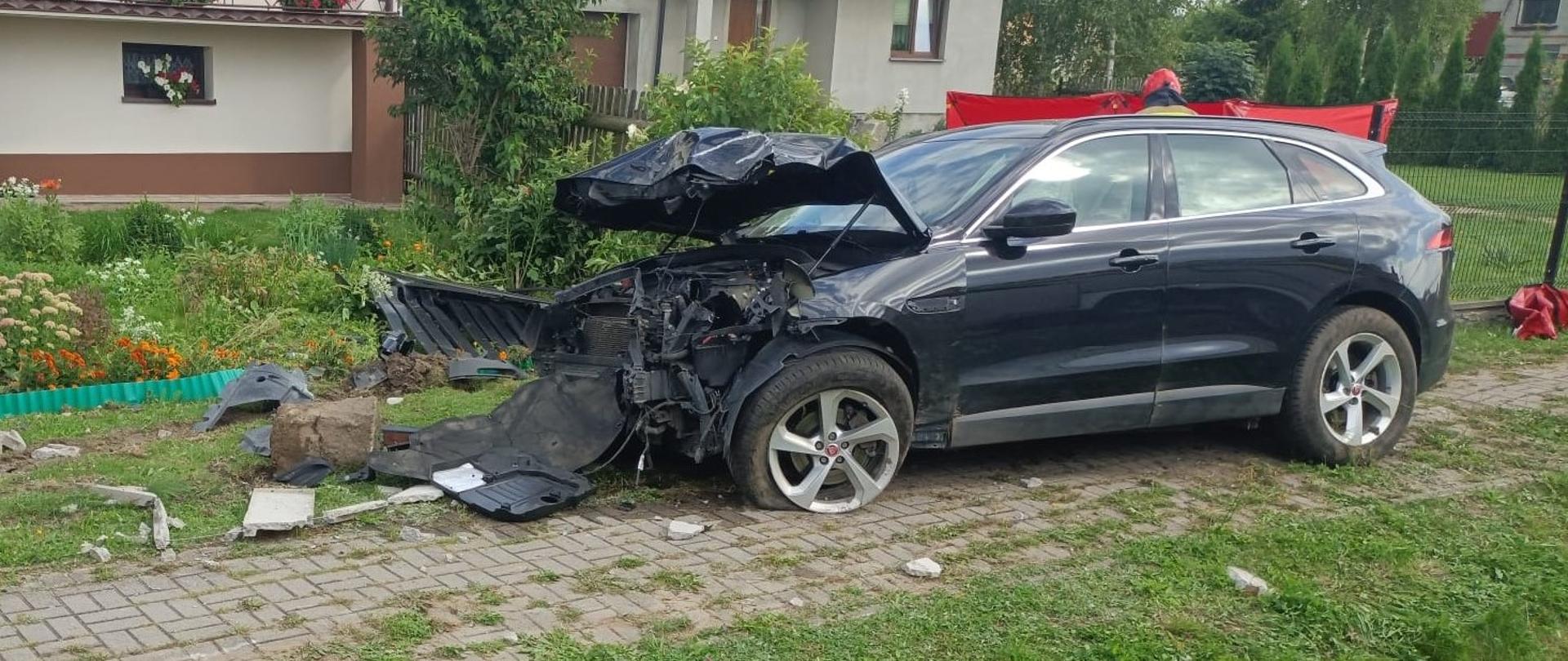 
Zdjęcie przedstawia czarny samochód osobowy, SUV, który uległ poważnemu wypadkowi i stoi na poboczu obok wybrukowanej ścieżki. Przód pojazdu jest całkowicie zniszczony, maska jest mocno wgnieciona i podniesiona, a zderzak i inne elementy karoserii są rozerwane i leżą na ziemi. Przednie lewe koło jest uszkodzone, z widocznym brakującym fragmentem opony. Samochód znajduje się na trawniku, w tle widać domy mieszkalne z balkonami, a także bujną roślinność i kwiaty. W tle po prawej stronie widać fragment czerwonego obiektu, prawdopodobnie należącego do służb ratowniczych. Niebo jest pochmurne.