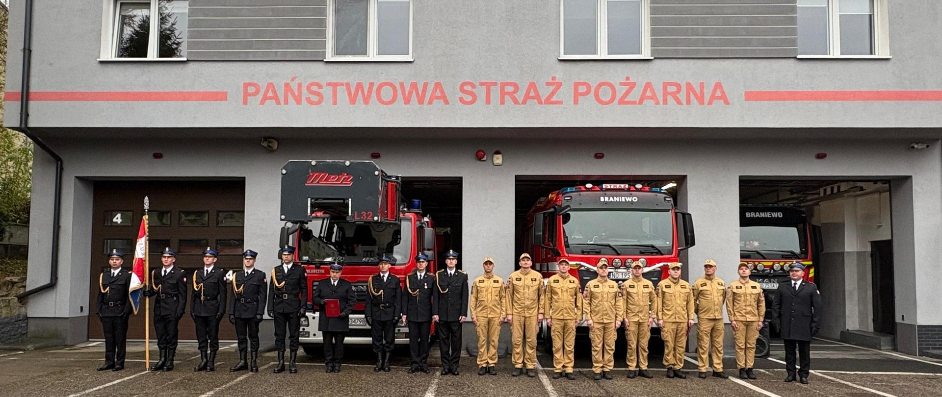 Na placu przed komendą stoi grupa strażaków, strażacy z lewej w mundurach galowych, na skraju poczet sztandarowy a z prawej w jasnych mundurach koszarowych. W tle szary budynek z napisem Państwowa Straż Pożarna.