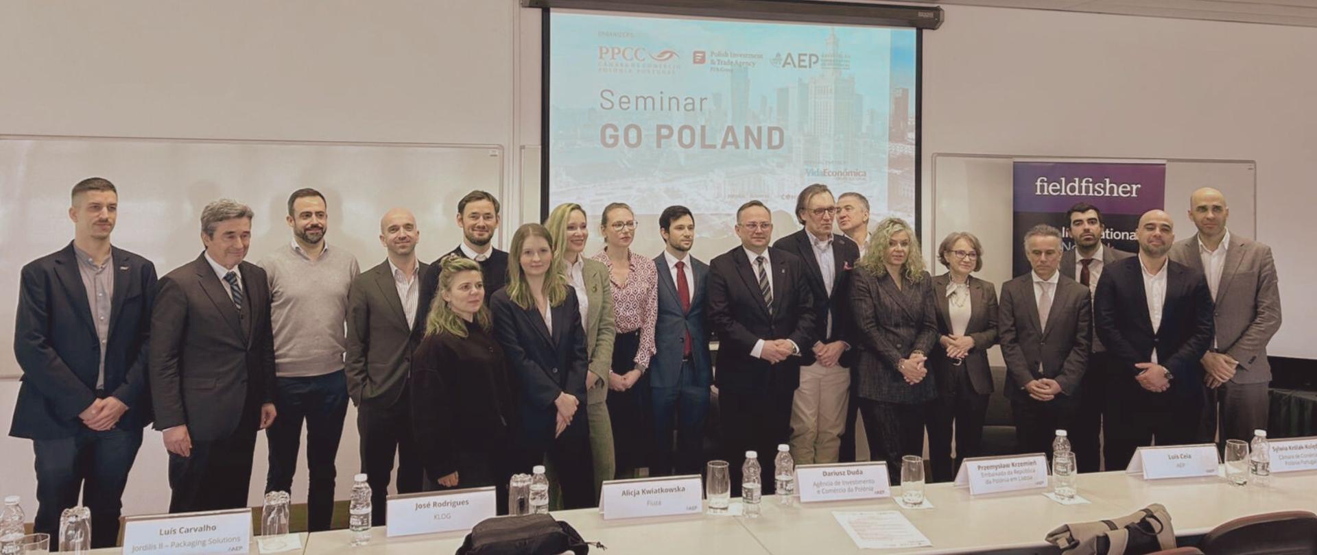 Grupa uczestników konferencji stoi za długim biurkiem pozując do zdjęcia. Za nimi, na dużym ekranie, widnieje napis o treści "Seminar Go Poland" oraz logotypy instytucji.