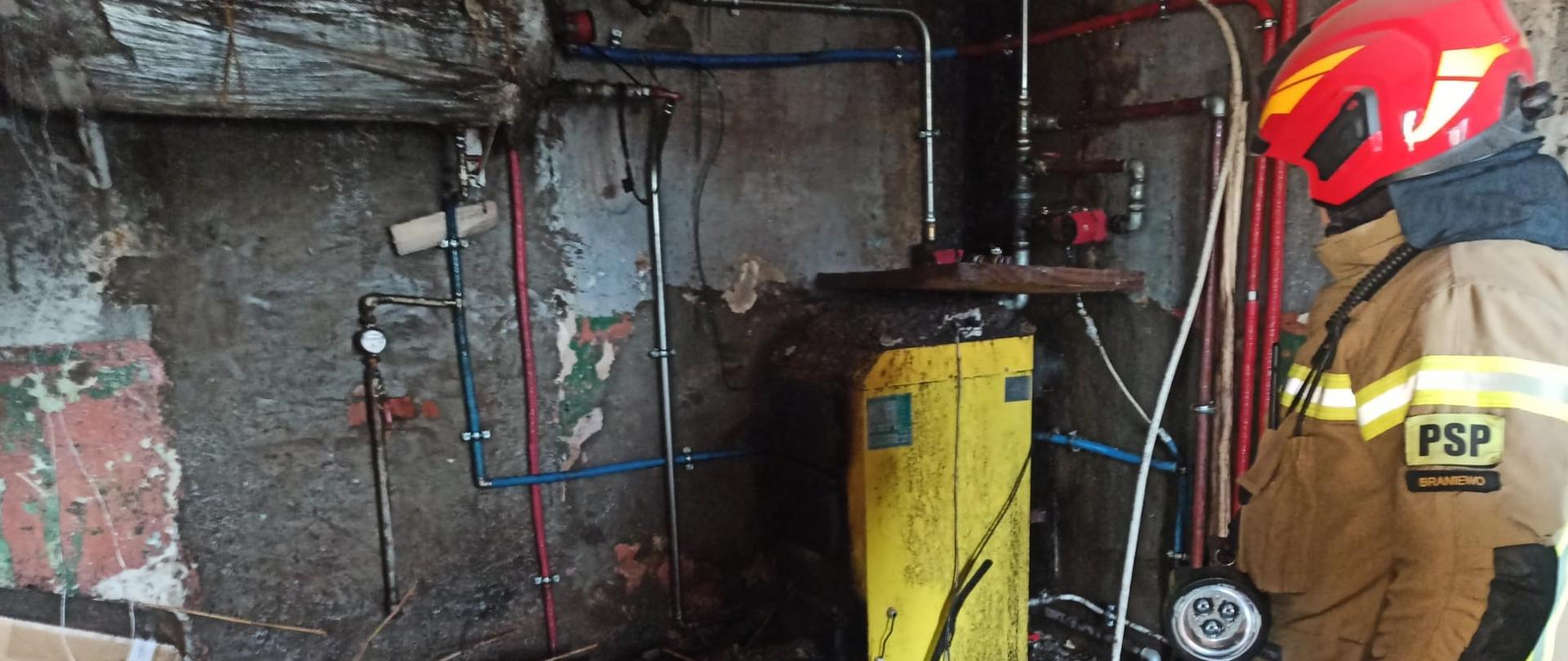Na zdjęciu pomieszczenie po pożarze. Czarne ściany. Z prawej strony stoi strażak w mundurze i czerwonym hełmie, w rogu żółty piec.