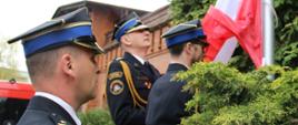 Flagowy i asystujący wciągają flagę na maszt. Obok stoi dowódca pocztu. W tle budynek i zieleń z trawnika.