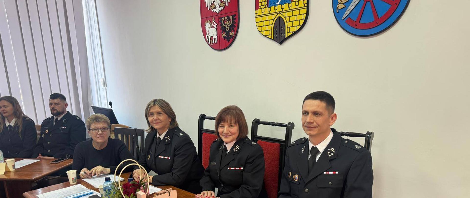 Trwa spotkanie. Z stołem siedzą strażacy i strażaczki w mundurach galowych.