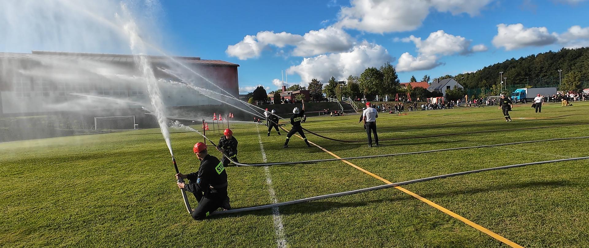 Ćwiczenie alarmowo-bojowe. Strażacy przekraczają linie z wężami strażackimi.