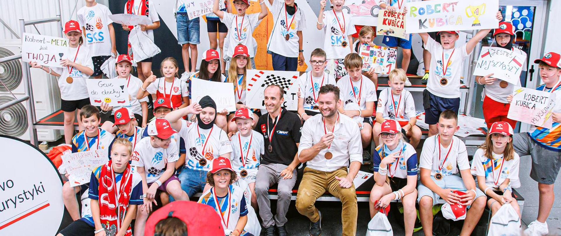 Robert Kubica i minister Kamil Bortniczuk siedzą na ławce a w raz z nimi dużo małych dzieci. Wszyscy są uśmiechnięci a niektóre z dzieci trzymają namalowane przez siebie plakaty z napisami: Robert Kubica, Jesteś najszybszy, Gaz do dechy i tym podobne. 