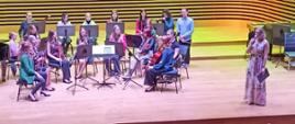 Muzycy orkiestry siedzą na krzesłach z instrumentami, obok stoi dyrygent, po prawej stronie kobieta mówi do mikrofonu.