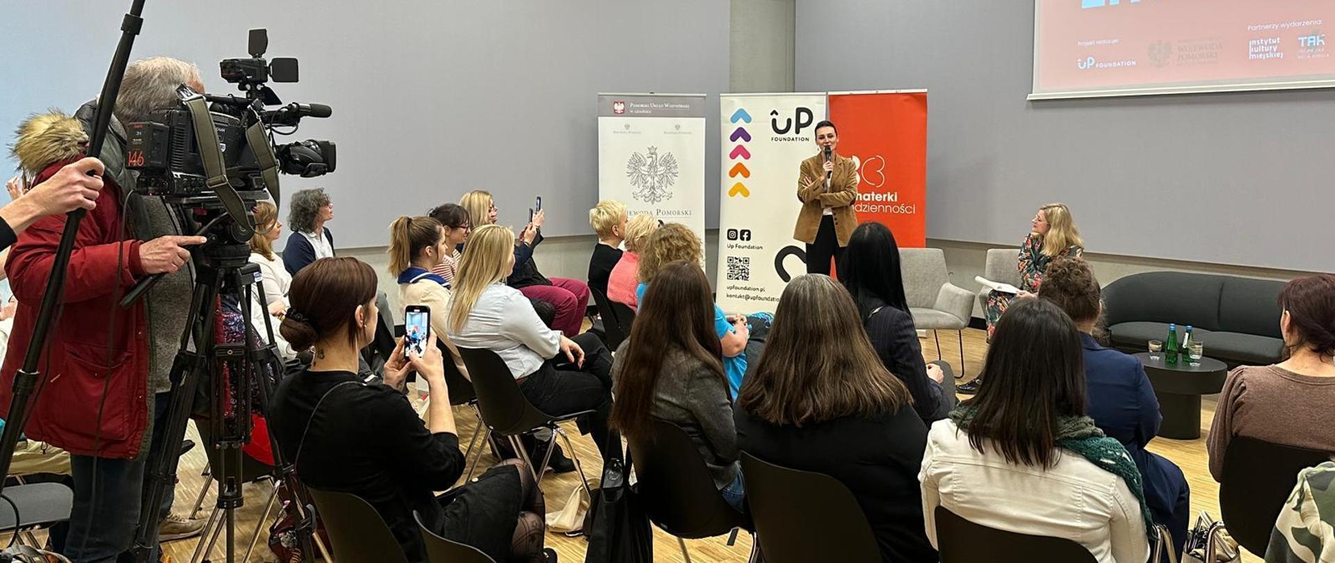 Sala, spotkanie z liderkami społecznymi. Przemawia wojewoda, uczestniczki siedzą na krzesłach, obok nich kamera telewizyjna