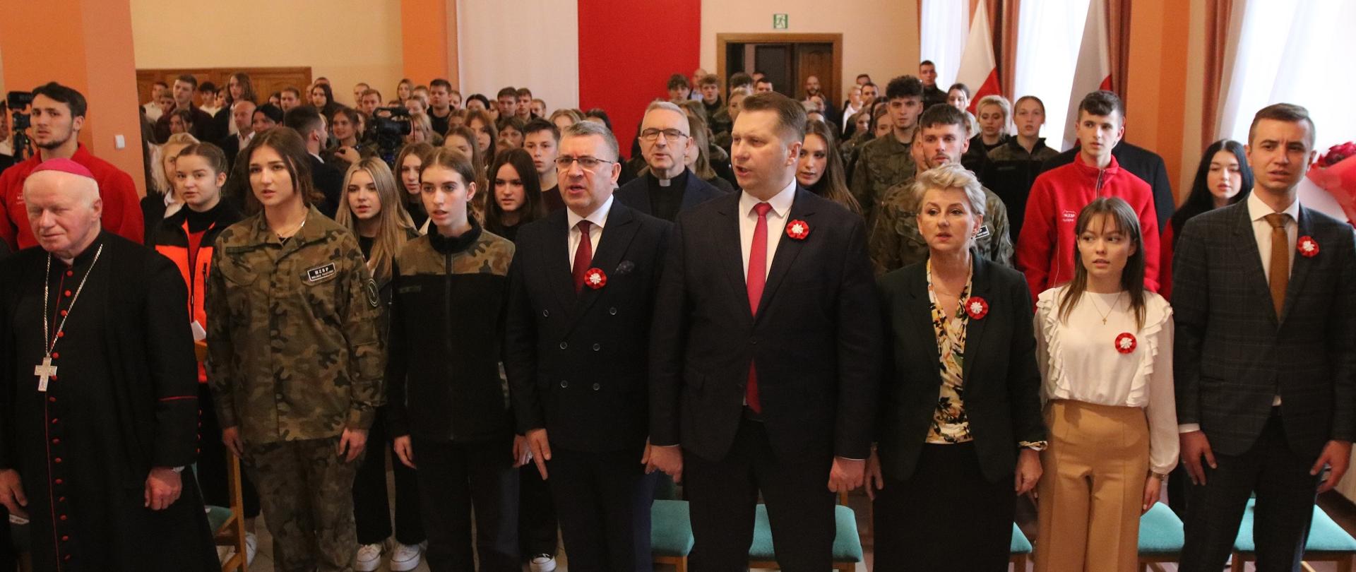 Duża grupa ludzi, wśród nim minister Czarnek, stoi w sali i śpiewa.