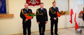 Na zdjęciu widzimy 3 funkcjonariuszy stojących na tle godła państwowego dwóch z nich trzyma w rękach kwiaty. Na zdjęciu przedstawiony jest Komendant Miejski PSP w Tarnowie wraz ze swoim nowo powołanym zastępcą oraz dotychczasowy zastępca przechodzący na emeryturę. Oficerowie ubrani są umundurowanie galowe. 