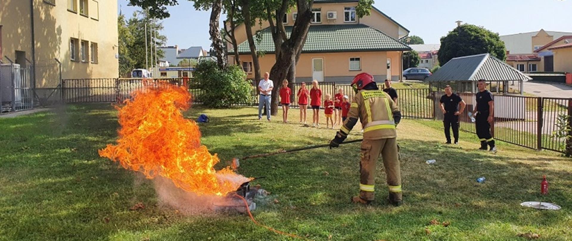Zdjęcie obrazuje strażaka gaszącego pozorowany pożar w tle strażacy i członkowie MDP OSP