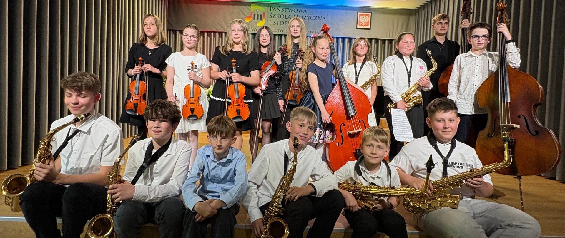 Na zdjęciu znajduje się grupa dzieci z instrumentami - skrzypcami, saksofonami i kontrabasami. W tle logo szkoły i godło, na szarych kotarach.