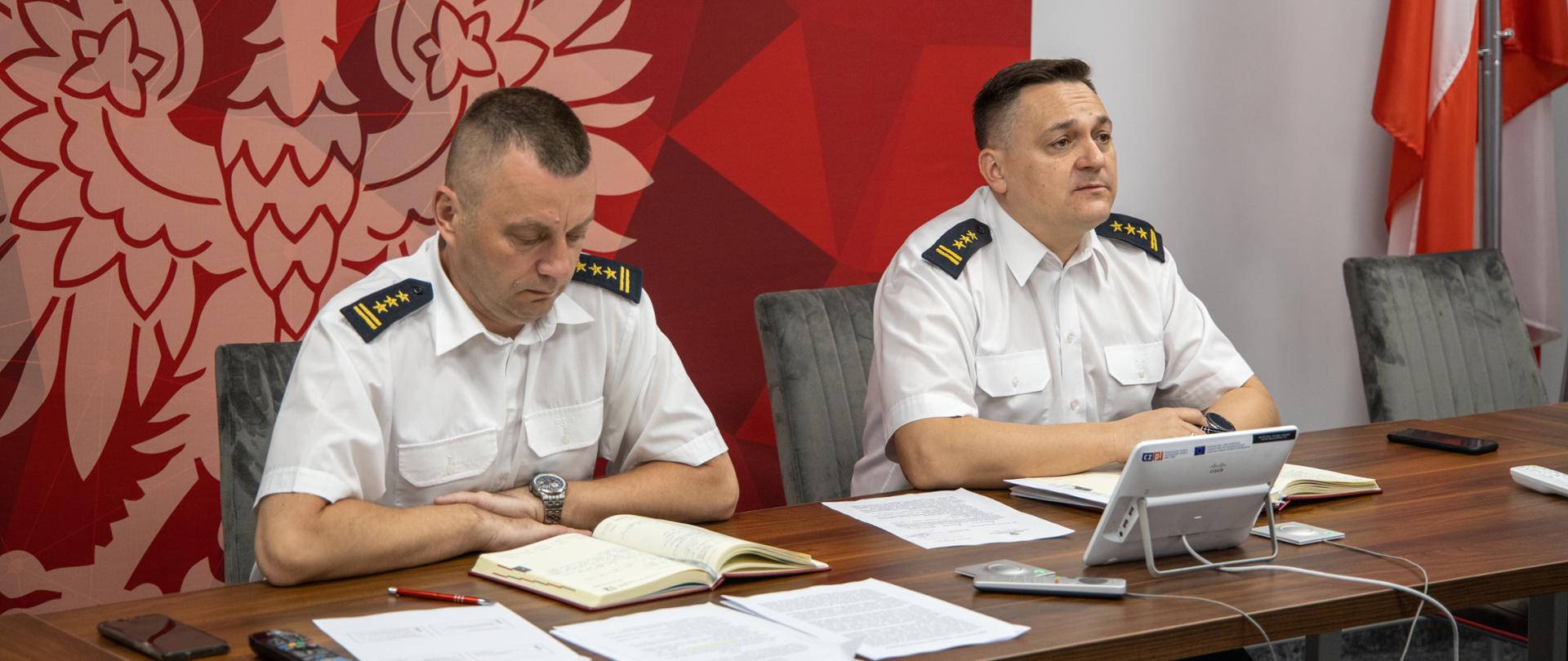 Zdjęcie przedstawia dwóch strażaków w ubraniu wyjściowym letnim (koszula letnia) siedzących przed stołem podczas prowadzonej wideokonferencji. W tle ścianka z godłem Polski oraz flag Rzeczypospolitej Polskiej.
Na zdjęciu znajduje się od prawej st. bryg. Krzysztof Kędryk Opolski Komendant Wojewódzki PSP oraz st. bryg. Arkadiusz Kuśmierski Zastępca Opolskiego Komendanta Wojewódzkiego PSP.
