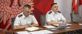 Zdjęcie przedstawia dwóch strażaków w ubraniu wyjściowym letnim (koszula letnia) siedzących przed stołem podczas prowadzonej wideokonferencji. W tle ścianka z godłem Polski oraz flag Rzeczypospolitej Polskiej.
Na zdjęciu znajduje się od prawej st. bryg. Krzysztof Kędryk Opolski Komendant Wojewódzki PSP oraz st. bryg. Arkadiusz Kuśmierski Zastępca Opolskiego Komendanta Wojewódzkiego PSP.
