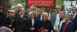 Na zdjęciu widoczna jest grupa osób zgromadzonych w lesie podczas oficjalnego wydarzenia. Uczestnicy stoją przed tablicą informacyjną rezerwatu przyrody „Lasy Sopockie”, a jedna z osób trzyma okolicznościową teczkę z dokumentem. Wśród zebranych znajdują się przedstawiciele różnych instytucji i organizacji, a na pierwszym planie widać sprzęt wykorzystywany do rejestrowania przebiegu wydarzenia. W tle rozciąga się naturalny las z wysokimi drzewami i gęstą roślinnością.