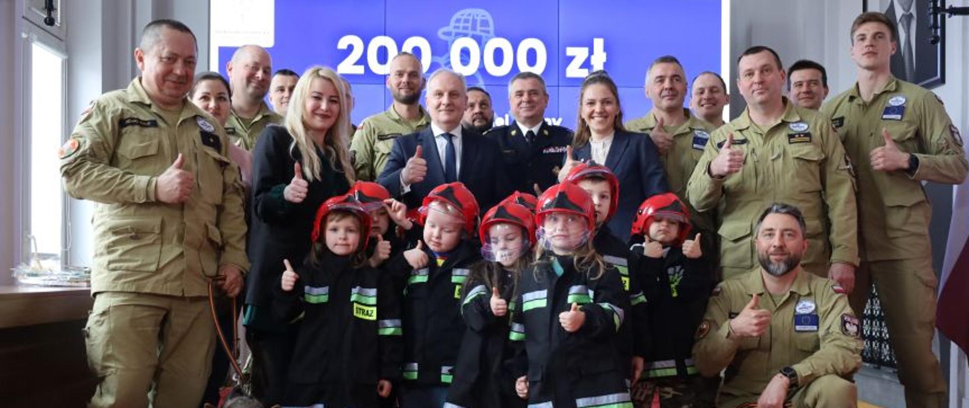 Strażacy dzieci oraz osoby cywilne stoją obok siebie wspólnie z dwoma psami za nimi na ekranie rzutnika wyświetlana jest kwota dwieście tysięcy złotych.