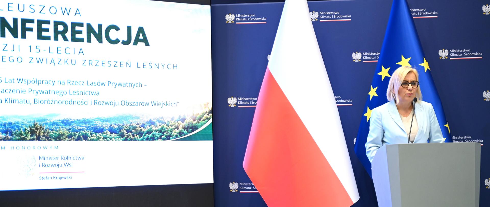Osoba przemawiająca przy mównicy podczas konferencji. W tle flagi Polski i Unii Europejskiej oraz ekran z nazwą konferencji.