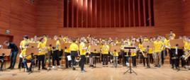 Blisko setka dzieci w żółtych koszulkach trzymających waltornie stoi na scenie w kilku rzędach przy pulpitach
