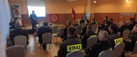 Na zdjęciu w centralnej części stoi starszy brygadier Rafał Rutyna w umundurowaniu wyjściowym na tle ekranu z wyświetloną prezentacją . Po prawej stronie widoczny znak Związku Ochotniczych Straży Pożarnych a następnie widoczne sztandary. W lewej stronie zdjęcia widoczny baner z wizerunkiem świętego Floriana. Przed nim druhowie w umundurowaniu koszarowym siedzą na krzesłach w odstępach około 1,5 m. 
