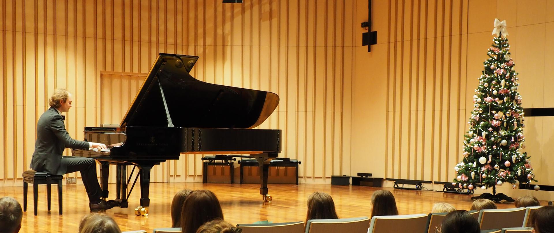 Pianista gra na fortepianie na scenie w sali koncertowej, na pierwszym planie publiczność, po prawej stronie sceny choinka bożonarodzeniowa.