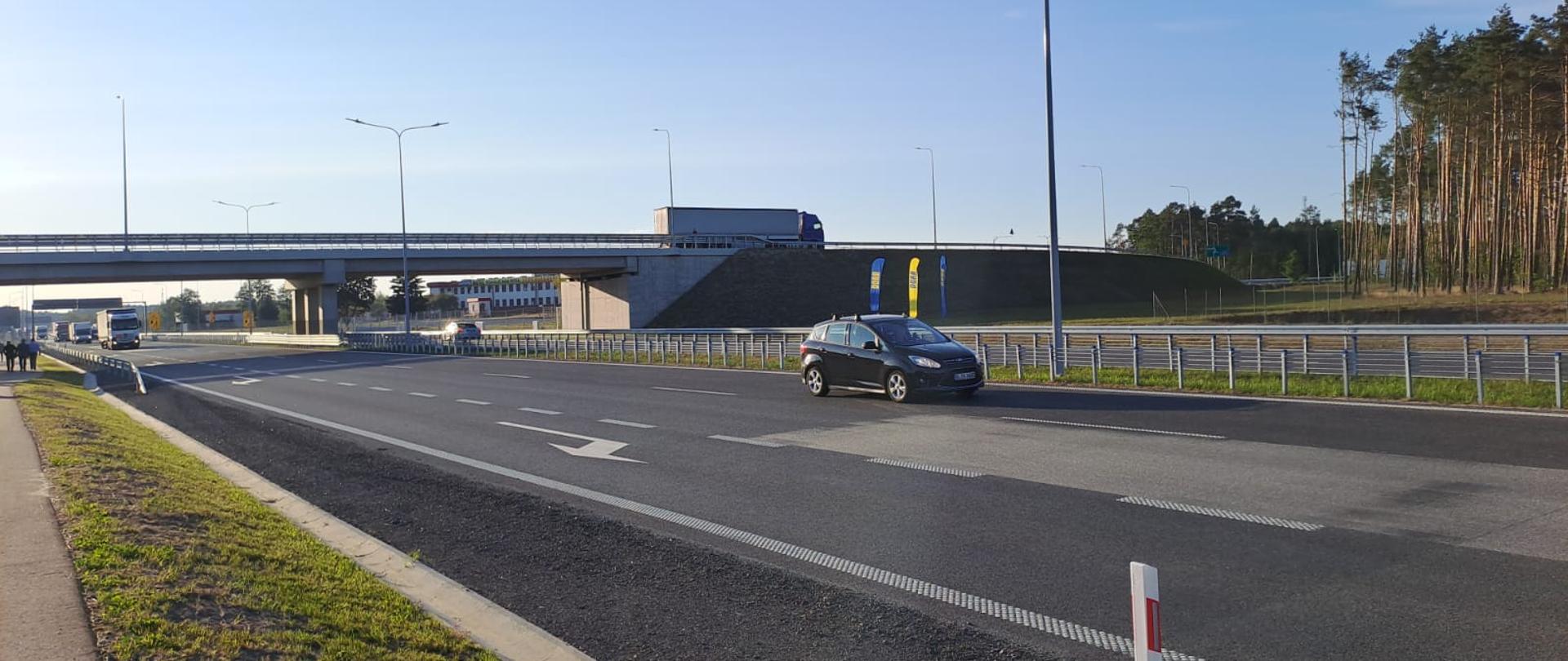 Samochody na dwupasmowej drodze. Na drugim planie wiadukt.