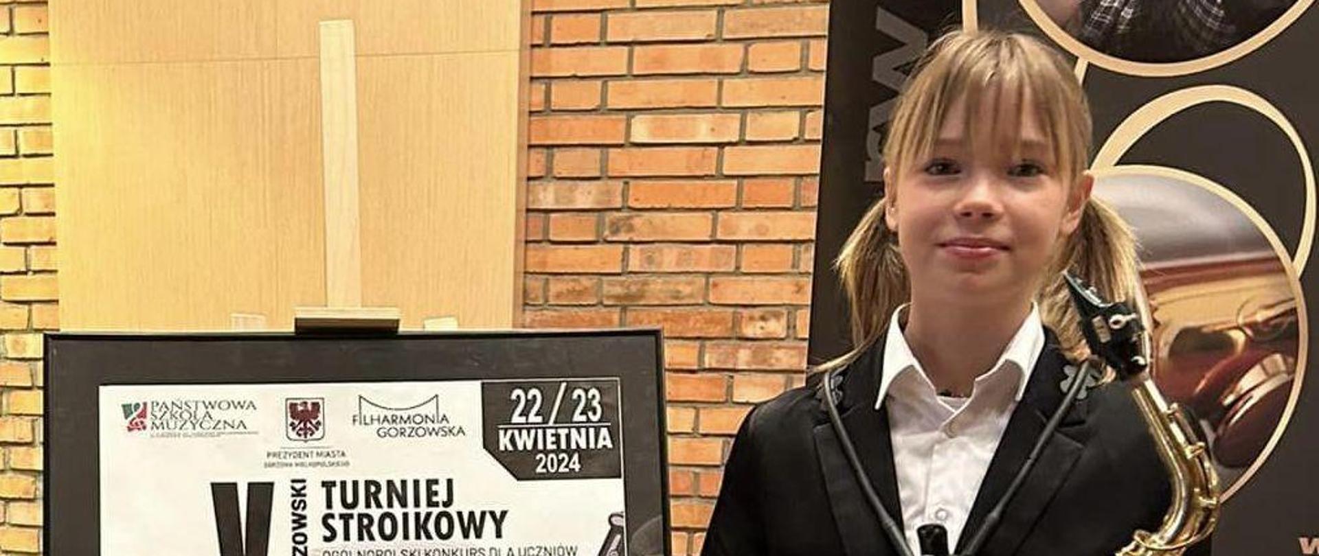 Uczennica Laura Nowak stoi trzymając saksofon. Obok plakat reklamujący V Gorzowski Turniej Stroikowy.