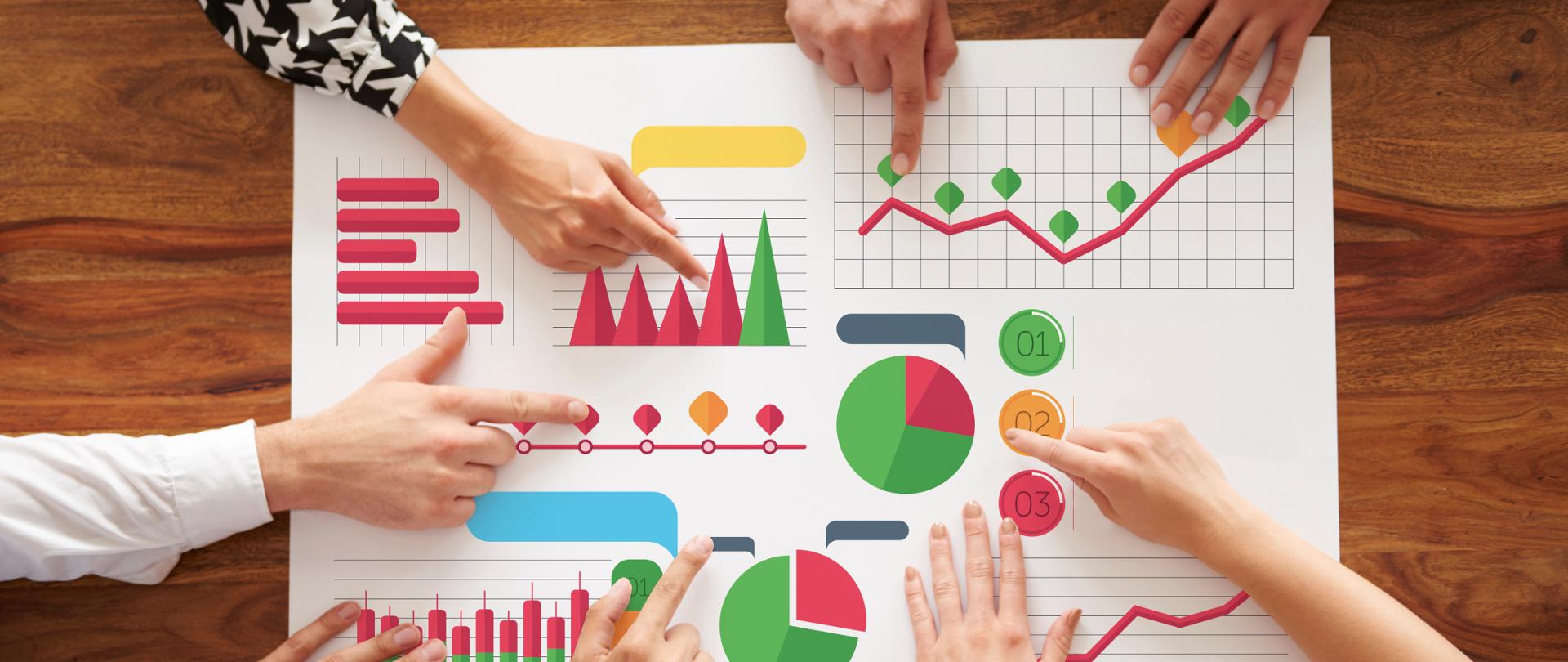 Zdjęcie białej planszy z kolorowymi wykresami i diagramami prezentującymi dane. Naokoło ludzkie dłonie wskazujące na poszczególne wykresy.