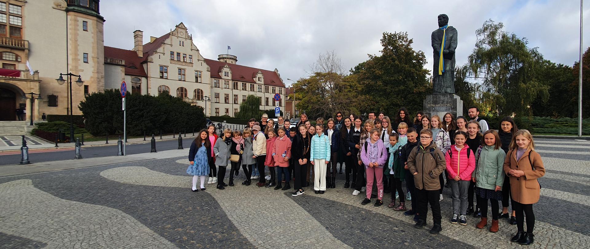 Uczniowie, rodzice i nauczyciele stojący na placu przed pomnikiem Adama Mickiewicza w Poznaniu.