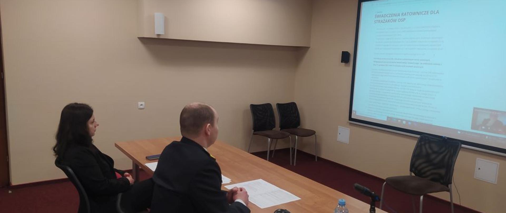 Przy brązowym stoliku siedzi mężczyzna i kobieta w ciemnych ubraniach odwróceni tyłem. Przed nimi ekran z wyświetlonym drobnym drukiem tekstem.