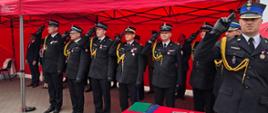 Strażacy w galowych mundurach salutują podczas oficjalnej części uroczystości pod czerwonym namiotem.
