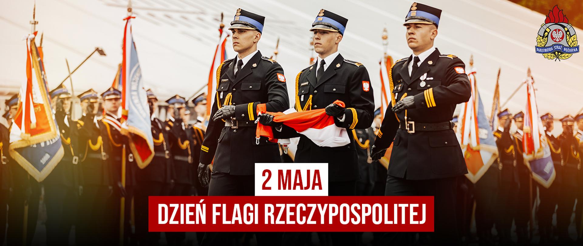 Strażacy PSP w umundurowaniu galowym maszerują w szyku, niosąc flagę Polski. W tle widoczne sztandary i formacja honorowa. Na grafice napis: „2 maja – Dzień Flagi Rzeczypospolitej”.