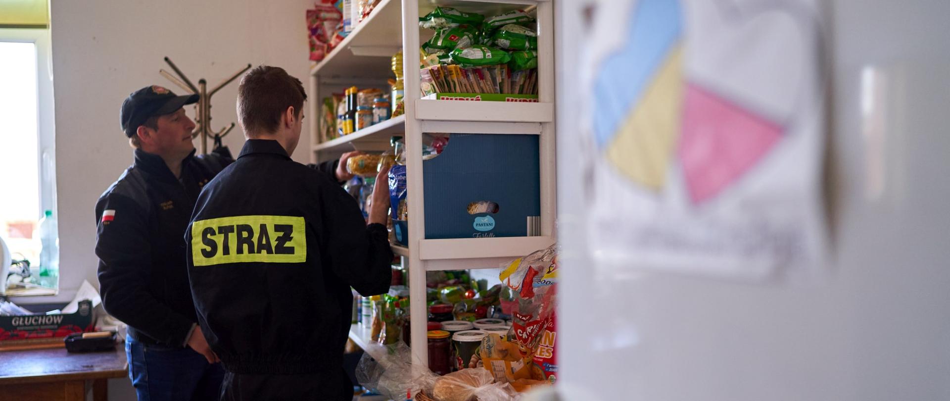 Zdjęcie przedstawia pomieszczenie.Po prawej stronie drzwi lodówki z naklejonym rysunkiem flagi Polski i Ukrainy w kształcie serc. Dalej po prawej stronie półki z żywonością, którą segreguje dwóch strażaków. Przy lewej krawędzi zdjęcia fragment okna.