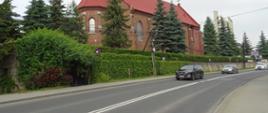 Zdjęcie przedstawia drogę jednopasmową po jednym pasie w każdym kierunku. W tle widoczny kościół z cegły i czerwoną dachówką.