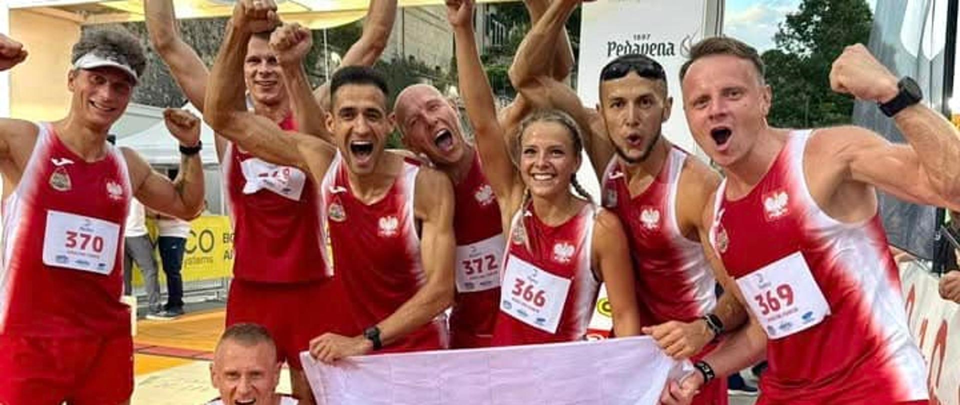 Kilkunastu strażaków w strojach sportowych - stoją w grupie i z podniesionymi rękoma manifestują zwycięstwo. Z przodu rozłożyli flagę biało-czerwoną. Za nimi transparenty, brama mety.